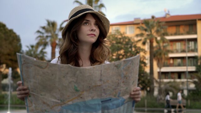 Young caucasian woman in hat holding a paper map on background of blurred hotel and palms. Woman long hair holding a map with eyes up, tourist in french city