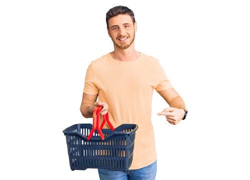 Handsome young man with bear holding supermarket shopping basket smiling happy pointing with hand and finger