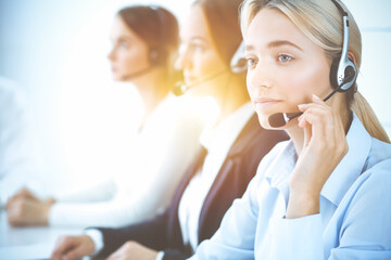 Cheerful smiling business woman with headphones consulting clients. Group of diverse phone operators at work in sunny office.Call center and business people concept