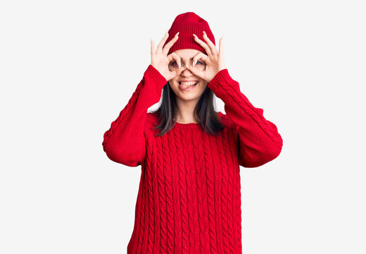 Young beautiful girl wearing sweater and wool cap doing ok gesture like binoculars sticking tongue out, eyes looking through fingers. crazy expression.