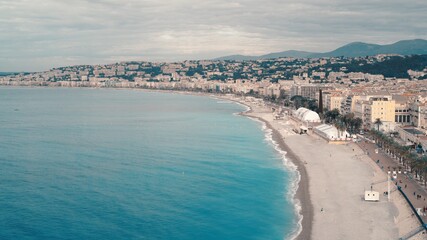 Fototapeta premium South city with a bright coastline in Nice, France, houses near a big ocean. Cloudy day, drone shot of long extended shoreline, cloudy windy day, sandy beach mountains on background
