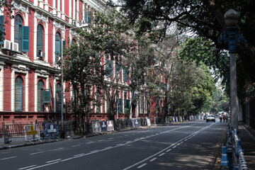 Fototapeta na wymiar Kolkata, India - February 1, 2020: Three unidentified people walks on the street with two passing cars next to a red house facade on February 1, 2020 in Kolkata, India