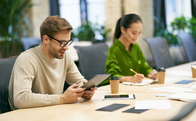 Working together. Young smiling caucasian man wearing eyeglasses using digital tablet while sitting at desk with his female colleague in the modern coworking space