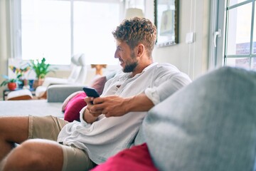 Fototapeta premium Handsome caucasian man smiling happy sitting on the sofa at home using smartphone
