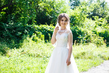 Fototapeta na wymiar Happy bride in a long white wedding dress and veil in a green park on nature. Wedding image of a young girl, women's makeup and hairstyle. Marriage concept