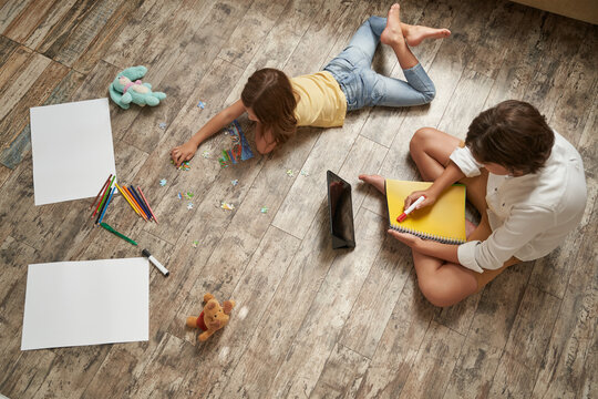 Brother and sister lying on the wooden floor at home and spending time together. Little girl playing with puzzles, boy using digital tablet and writing something in notebook