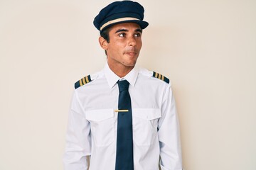 Young hispanic man wearing airplane pilot uniform smiling looking to the side and staring away thinking.