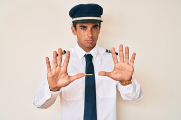 Young hispanic man wearing airplane pilot uniform moving away hands palms showing refusal and denial with afraid and disgusting expression. stop and forbidden.