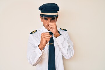 Young hispanic man wearing airplane pilot uniform ready to fight with fist defense gesture, angry and upset face, afraid of problem