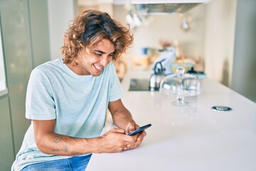Young hispanic man smiling happy using smartphone sitting on the table at home
