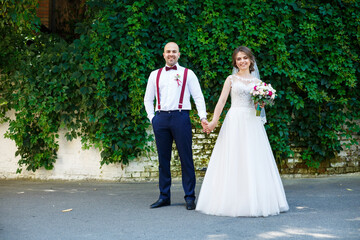 Beautiful couple bride in a white dress with a bouquet while the groom with suspenders and bow tie holding hands. Against the background of a wall with green leaves. Happy couple. Marriage concept.