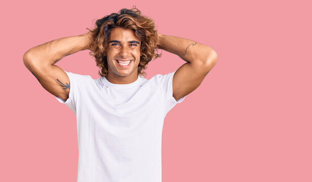 Young hispanic man wearing casual white tshirt relaxing and stretching, arms and hands behind head and neck smiling happy