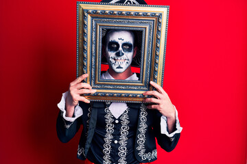 Young man wearing mexican day of the dead costume holding empty frame looking positive and happy standing and smiling with a confident smile showing teeth