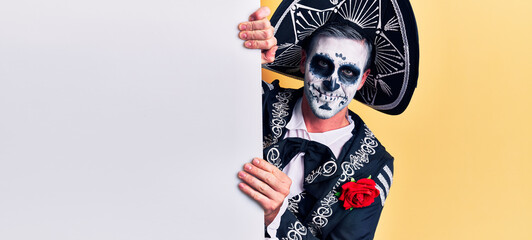 Young man wearing mexican day of the dead costume holding blank empty banner looking positive and happy standing and smiling with a confident smile showing teeth
