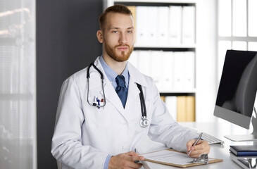 Friendly red-bearded doctor sitting and writing at clipboard in clinic. Perfect medical service in hospital. Medicine concept