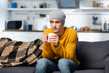 cold man in knitted sweater and hat looking at camera while holding cup of warm tea in kitchen