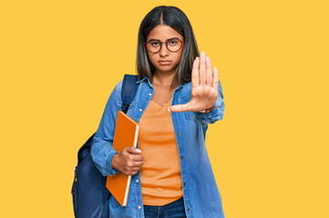 Young latin girl wearing student backpack and holding books doing stop sing with palm of the hand. warning expression with negative and serious gesture on the face.