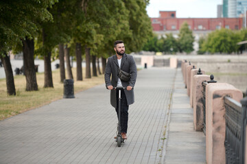 Contemporary young bearded businessman in coat riding on electric scooter