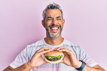 Middle age grey-haired man eating a tasty classic burger smiling and laughing hard out loud because funny crazy joke.