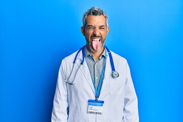 Middle age grey-haired man wearing doctor uniform and stethoscope sticking tongue out happy with funny expression. emotion concept.