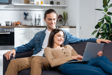 young man sitting on sofa in kitchen near joyful woman using laptop