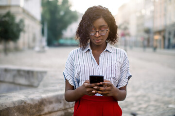 Close up horizontal portrait of attractive young african woman, wearing red pants and striped shirt, reading or typing text message on mobile phone at old city street. People, technologies concept