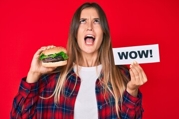 Beautiful caucasian woman eating a tasty classic burger holding wow text angry and mad screaming frustrated and furious, shouting with anger looking up.