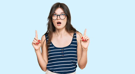 Young beautiful caucasian girl wearing casual clothes and glasses amazed and surprised looking up and pointing with fingers and raised arms.