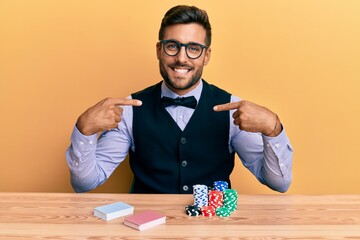 Handsome hispanic croupier man sitting on the table with poker chips and cards looking confident...
