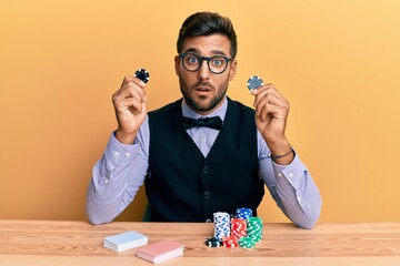 Handsome hispanic croupier man sitting on the table holding casino chips in shock face, looking...