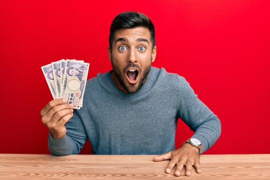 Handsome hispanic man holding japanese yen banknotes scared and amazed with open mouth for surprise, disbelief face