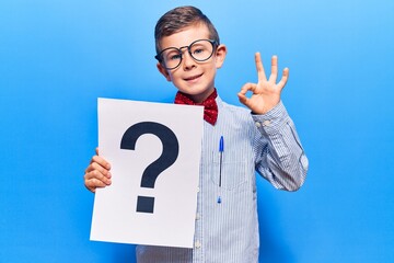 Cute blond kid wearing nerd bow tie and glasses holding question mark doing ok sign with fingers, smiling friendly gesturing excellent symbol
