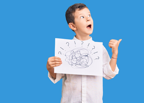 Cute blond kid holding scribble draw pointing thumb up to the side smiling happy with open mouth