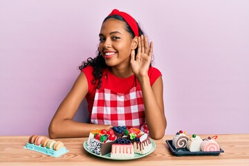 Young african american girl wearing baker uniform sitting on the table with sweets smiling with hand over ear listening and hearing to rumor or gossip. deafness concept.
