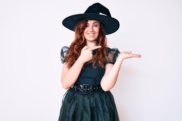 Young beautiful woman wearing witch halloween costume amazed and smiling to the camera while presenting with hand and pointing with finger.