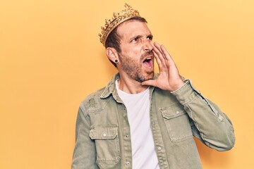 Young handsome man wearing king crown shouting and screaming loud to side with hand on mouth. communication concept.