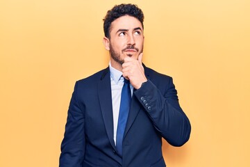 Young hispanic man wearing suit with hand on chin thinking about question, pensive expression. smiling with thoughtful face. doubt concept.