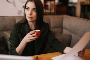Focused young business woman studying paper documents in cafe sitting at table with laptop. Lady enjoys delicious coffee while working. Student girl sitting in cafe with cup of coffee.