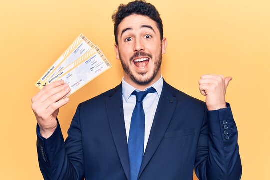 Young hispanic man wearing suit holding boarding pass pointing thumb up to the side smiling happy with open mouth