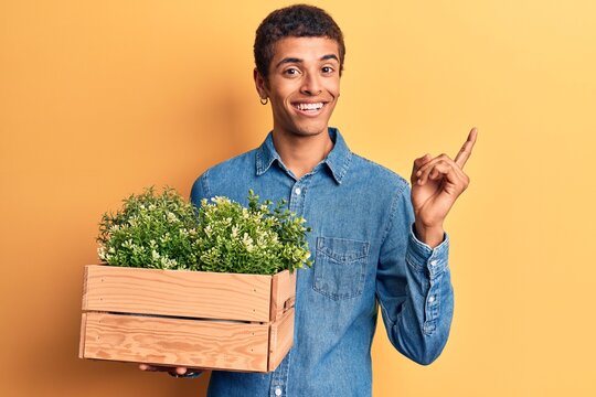 Young african amercian man holding wooden pot with plant smiling happy pointing with hand and finger to the side