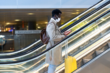 Afro-American traveler man with yellow suitcase stands on escalator in airport terminal, wear face medical mask to protect yourself from contact with flu virus, pandemic covid-19. New normal concept 