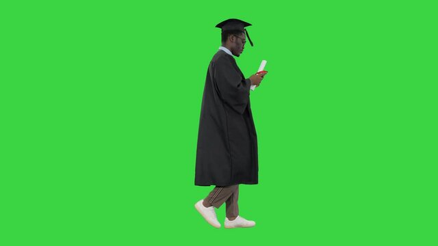 African american male student in graduation robe texting on the phone while walking with his diploma on a Green Screen, Chroma Key.