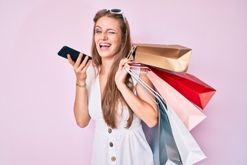 Young blonde girl holding shopping bags talking on the smartphone winking looking at the camera...