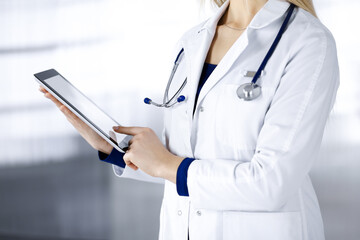 Unknown woman-doctor is holding a tablet computer in her hands, while standing in a clinic. Female physician with a stethoscope in her office, close-up. Perfect medical service in a hospital. Medicine