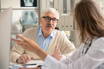 Senior patient in eyeglasses looking at doctor pointing at computer screen