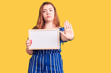 Naklejka premium Young beautiful redhead woman holding empty white chalkboard with open hand doing stop sign with serious and confident expression, defense gesture