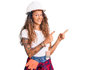 Young hispanic woman with tattoo wearing hardhat and builder clothes smiling and looking at the camera pointing with two hands and fingers to the side.