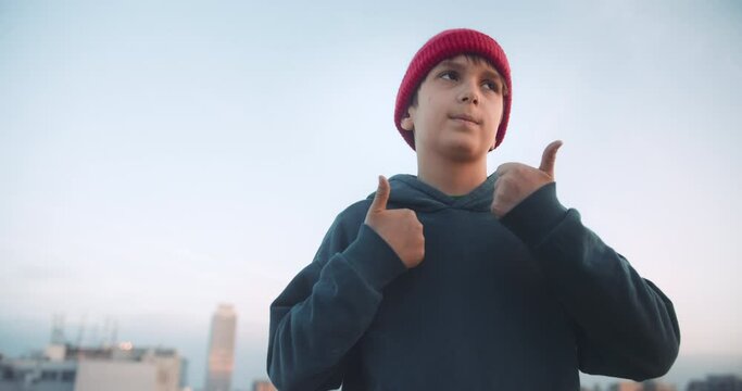 Handsome young boy dancing and have fun on rooftop at sunset. Wearing hoody and beanie red hat looking confident in urban city background.