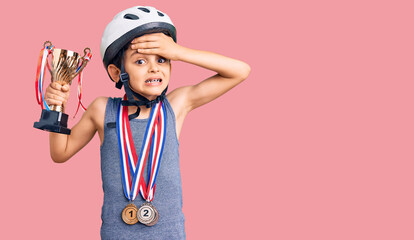 Little cute boy kid wearing bike helmet and winner medals holding winner trophy stressed and frustrated with hand on head, surprised and angry face