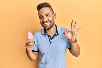 Handsome man with beard eating ice cream cone doing ok sign with fingers, smiling friendly...